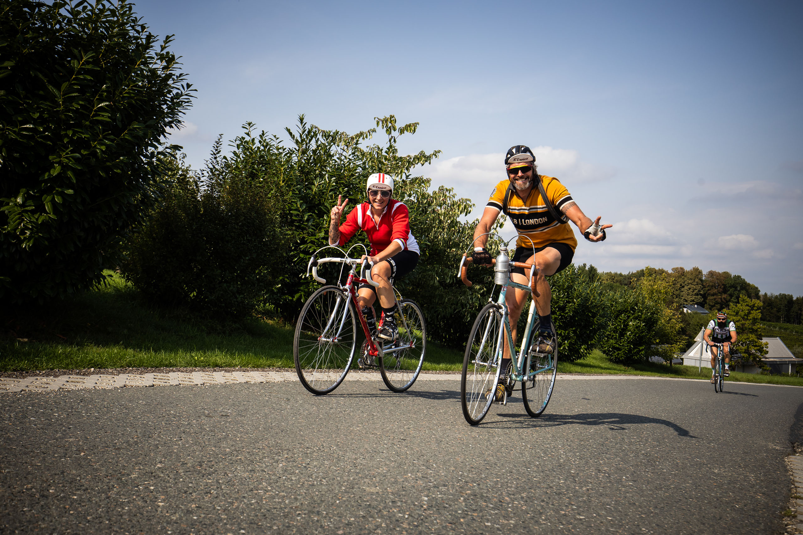 Radfahrer:innen im Vintage-Look fahren entlang einer Straße im Grünen bei Styroica - Vintage Radrundfahrt im Thermen- und Vulkanland. Radfahrer:innen im Vintage-Look auf alten Fahrrädern nehmen an der Styroica teil, der Vintage Radrundfahrt im Thermen- und Vulkanland.